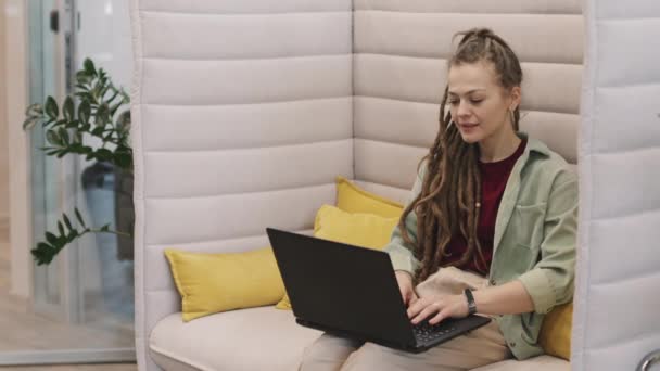 一个快乐的年轻女商人的慢吞吞的Pan 她带着恐惧的头发坐在现代初创办公室或合作空间的沙发上 在笔记本电脑上打字 — 图库视频影像