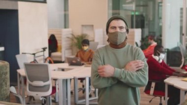 Şapkalı ve maskeli bir gencin kollarını açık ofiste çapraz pozu ya da ortak çalışma alanı.