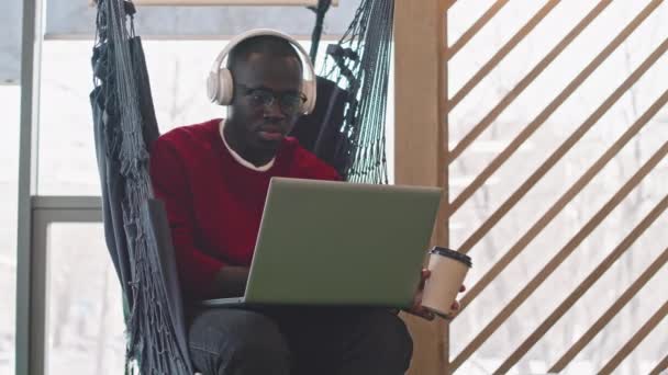 Powolne Ujęcie Afroamerykanina Słuchawkach Okularach Siedzącego Hamaku Nowoczesnej Przestrzeni Coworkingowej — Wideo stockowe