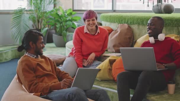 Pan拍摄的快乐的年轻人 他们带着笔记本电脑和快乐的女人坐在现代初创公司办公室的休息区里聊天 — 图库视频影像