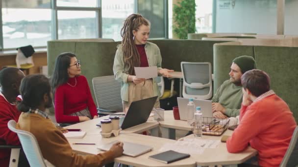 事務所のテーブルの周りに座っている若いビジネスマンのグループに文書を保持し 話すドレッドロックを持つ若い女性の遅いパン — ストック動画