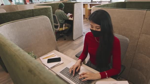 スタートアップオフィスやコワーキングスペースのブースに座って ラップトップで作業している顔マスクの若いアジアの女性の高角度Panショット — ストック動画
