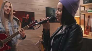 Siyah deri ceketli havalı bir kız ve mikrofonda bereyle şarkı söylerken, bayan meslektaşları müzik enstrümanları çalarken, retro-style stüdyoda konser provası yaparken...