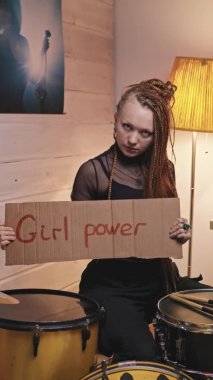 Rastalı genç gotik kadının dikey yavaş portresi. Kameraya bakıyor. Müzik stüdyosunda bateri setinde oturuyor ve elinde kız gücü yazan karton bir tabela var.