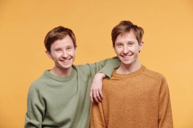 Genç ve mutlu bir adamın portresi. Günlük süveter giymiş. Kardeşinin omuzlarına yaslanıp turuncu arka plana yaslanıyor.