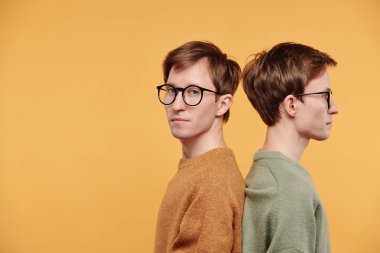 Hardal kazaklı ve gözlüklü genç adamın portresi. Turuncu arka planda kardeşiyle sırt sırta duruyormuş.