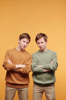 Çapraz kollarıyla turuncu arka planda dikilen sıradan süveter giyen asık suratlı gençlerin portresi.