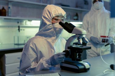 Koruyucu iş giyimi bilim adamı kimya laboratuarındaki masada mikroskopla numuneleri inceliyor.