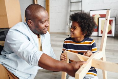 Mutlu genç Afrikalı adam ve sevimli küçük oğlu birbirlerine bakarken çocuk tahta sandalyeye çivi çakıyor.