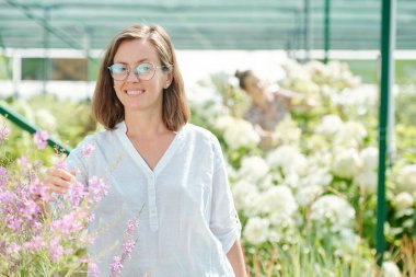 Beyaz keten gömlekli mutlu esmer kadın sera işçisi çiçeklerin arasında dikilirken dişlek bir gülümsemeyle sana bakıyor.