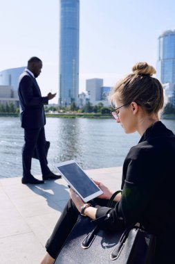 Resmi giyim ve gözlüklü genç iş kadını Nehir kenarında oturmuş Afrikalı işadamının önünde mesajlaşırken dijital tablet kullanıyor