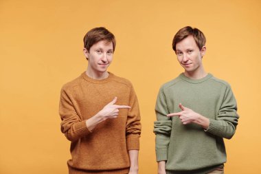 Süveter giyen, turuncu arka planda parmak tabancalarıyla birbirine doğrultulan içerik portresi.