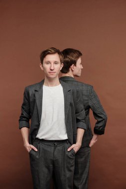 Gri takım elbiseli, kendine güvenen bir gencin portresi kardeşiyle sırt sırta, kahverengi arka plan.