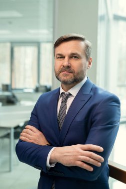 Koyu mavi takım elbiseli sakallı yöneticinin portresi kollarını kavuşturup ofis odasına yaslanmış duruyor.