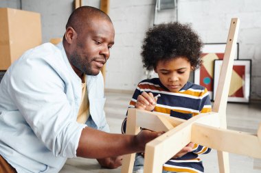 Genç Afrikalı bir adam, küçük sevimli oğluna tahta sandalyeyi nasıl monte edeceğini öğretiyor ve ona yardım ederken detaylarını da hallediyor.