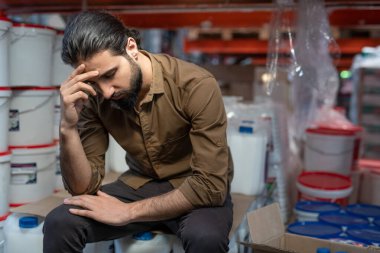 Puzzled young mixed race warehouse manager with beard sitting on canisters covered with cardboard and leaning head on hand clipart