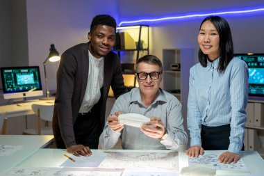 Olumlu, modern, çok ırklı araba tasarımcılarından oluşan bir ekip ofiste çizimler ve araba maketleriyle çalışıyor. Kameraya bakıyorlar.