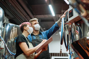 Çağdaş bira üretim fabrikasının iki genç bira üreticisi kontrol paneline bakarken, insanlar işleme fabrikasındaki çalışmalar sırasında verilere işaret ediyorlar.