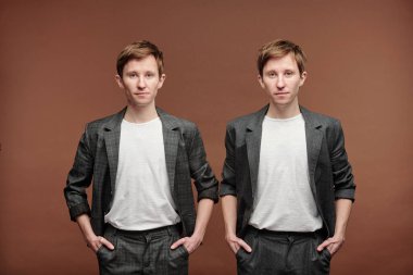 Şık takım elbiseli, beyaz tişörtlü, kahverengi arka planda pantolonun cebinde el ele tutuşan yakışıklı gençlerin portresi.