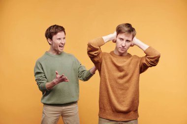 Bağıran kardeşi dinlemeyi reddedip kulaklarını kapatan sinirli genç adam, turuncu arka plan.