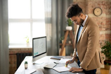 Bej ceketli, düşünceli genç ırk analisti masada duruyor ve çatı katında evrak okuyor.
