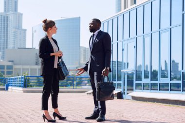 Kültürler arası takım elbiseli iki genç iş adamı güneşli bir günde şehir çevresindeki ofis binasına karşı çalışma anlarını tartışıyor.