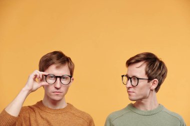 Zeytin rengi kazaklı genç adam, turuncu arka planda gözlük takan kardeşine bakıyor.