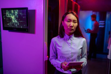 Sunucu odasındaki bilgisayar dolaplarına karşı elinde tabletle duran Asyalı ciddi bir kadın ağ yöneticisinin portresi