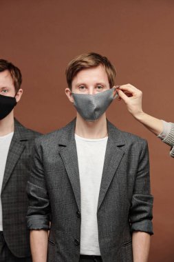 Gri takım elbiseli genç adamın kulağına maske takıyor. Kahverengi arka planda kardeşiyle ayakta duruyor.