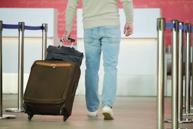 Mavi kotlu ve gri kazaklı bir erkek yolcunun havaalanındaki kapılardan geçerken bavulunu bavuluyla çekişi.