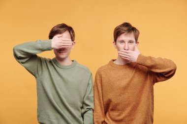 Süveter giymiş ergen kardeşlerin portresi turuncu arka planda duruyor ve gözleri ve ağzı kapalı akıllı maymunları canlandırıyor.