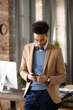 Yoğunlaşmış papyonlu genç siyah adam ofis masasında duruyor ve telefon mesajlarını kontrol ediyor.