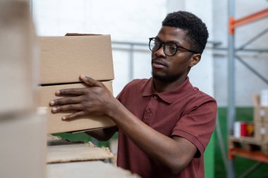 Polo tişörtlü, ciddi bir siyah işçi rafta duruyor ve depoda kutuları diziyor.
