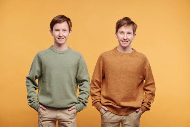 Elleri ceplerinde turuncu arka planda sıradan süveter giyen gülümseyen genç kızların portresi.