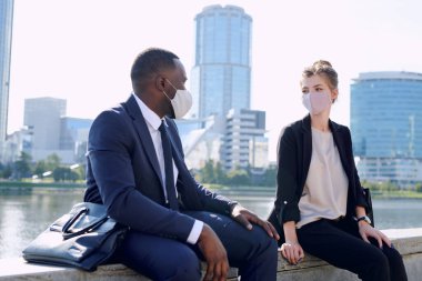 Formalite ve koruyucu maskeler içinde iki genç kültürler arası iş ortağı çalışma noktalarını tartışırken şehir ortamında oturuyorlar.