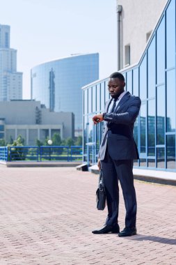Zarif koyu mavi takım elbiseli genç Afrikalı işadamı modern binalara ve güneşli bir günde parka karşı dururken kol saatine bakıyor.