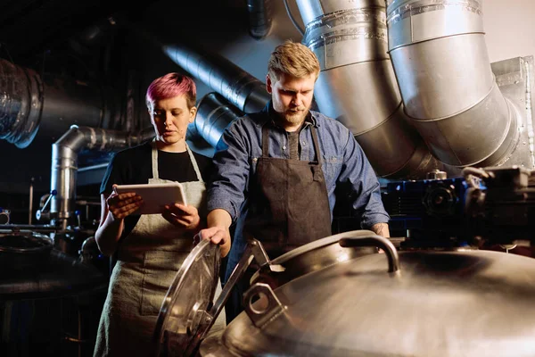 Young woman with digital tablet standing next to her male colleague opening lid of huge steel cistern during process of beer production