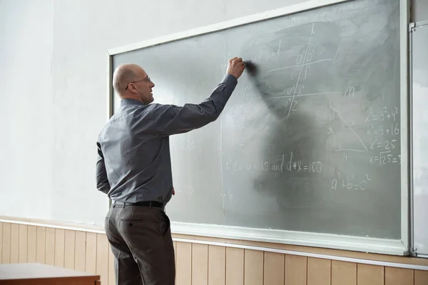 光秃秃的大学教授 穿着时髦的便服 站在黑板前 在讲解过程中写下代数方程 — 图库照片
