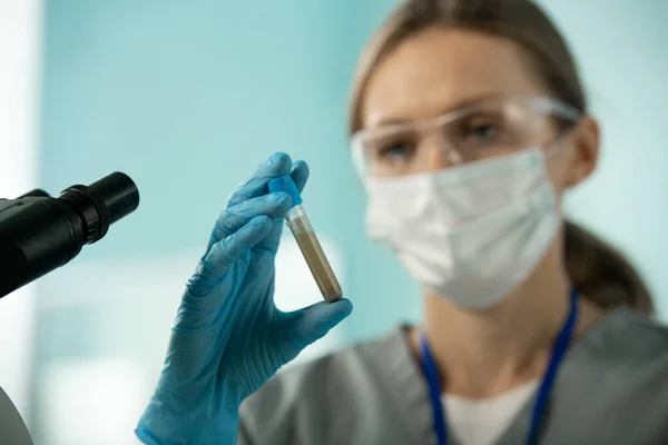 Close-up of young laboratory worker in mask and blue glove holding test tube of muddy liquid while working with samples and microscope