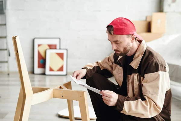 スタジオ 家やフラット内の木製の椅子を組み立てながら 紙の上の指示を見て若い家具アセンブリの専門家 — ストック写真