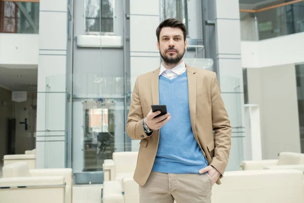 Portrait of content young bearded lawyer in blue sweater holding hand in pocket and checking smartphone in lobby