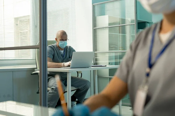 마스크와 수술복을 책상에 앉아서 컴퓨터로 보고서를 대머리 과학자 — 스톡 사진