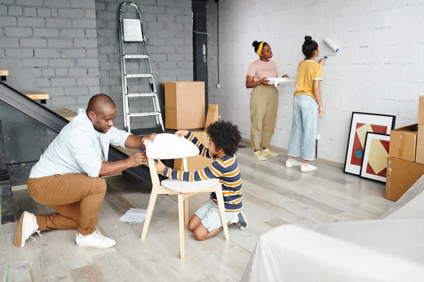 幼い父親と小さな息子が床の上に木製の椅子を組み立てる母親と娘に対して白い色のリビングルームの壁を描く — ストック写真