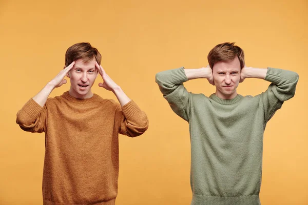 オレンジの背景を背景にセーターに悩まされた双子の肖像 そのうちの1つは 額の近くに他の手で耳をカバー — ストック写真