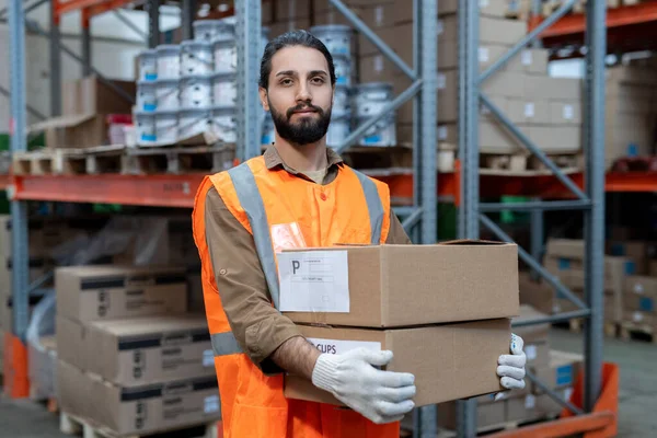 Portrait of content handsome mixed race mover in reflective vest carrying packed goods while preparing it for shipment