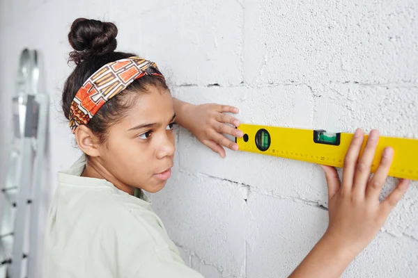 白い塗装レンガの壁に立っている深刻なアフリカの女性のティーンエイジャーと自宅の改修作業中にレベル測定ハンドツールを使用して — ストック写真