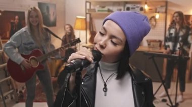 Havalı mor bereli havalı kızın yavaş yavaş yakın çekimi kameraya güvenle bakarken mikrofondaki bayan meslektaşlarıyla arka planda müzik aletleri çalarken rahat retro stüdyosunda