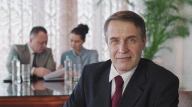 İş arkadaşları arka planda yeni projeyi tartışırken modern toplantı odasındaki konferans masasında kameraya gülümseyen orta yaşlı beyaz erkek yatırımcının yavaş belden yukarı portresi.