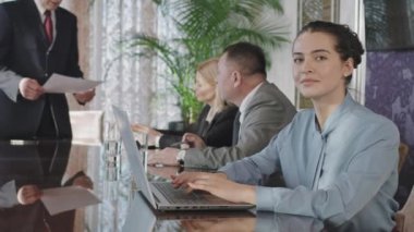 PAN waist-up slowmo portrait of young attractive business woman with laptop looking at camera sitting at negotiation table while her colleagues discussing new project in background