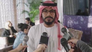 Geleneksel giysiler içindeki genç Arap işadamının belden yukarı çekimi, çok ırklı ortaklar ve yatırımcılarla yapılan resmi iş görüşmesi sırasında mikrofonla konuşan gazeteciler tarafından sorgulanıyor.